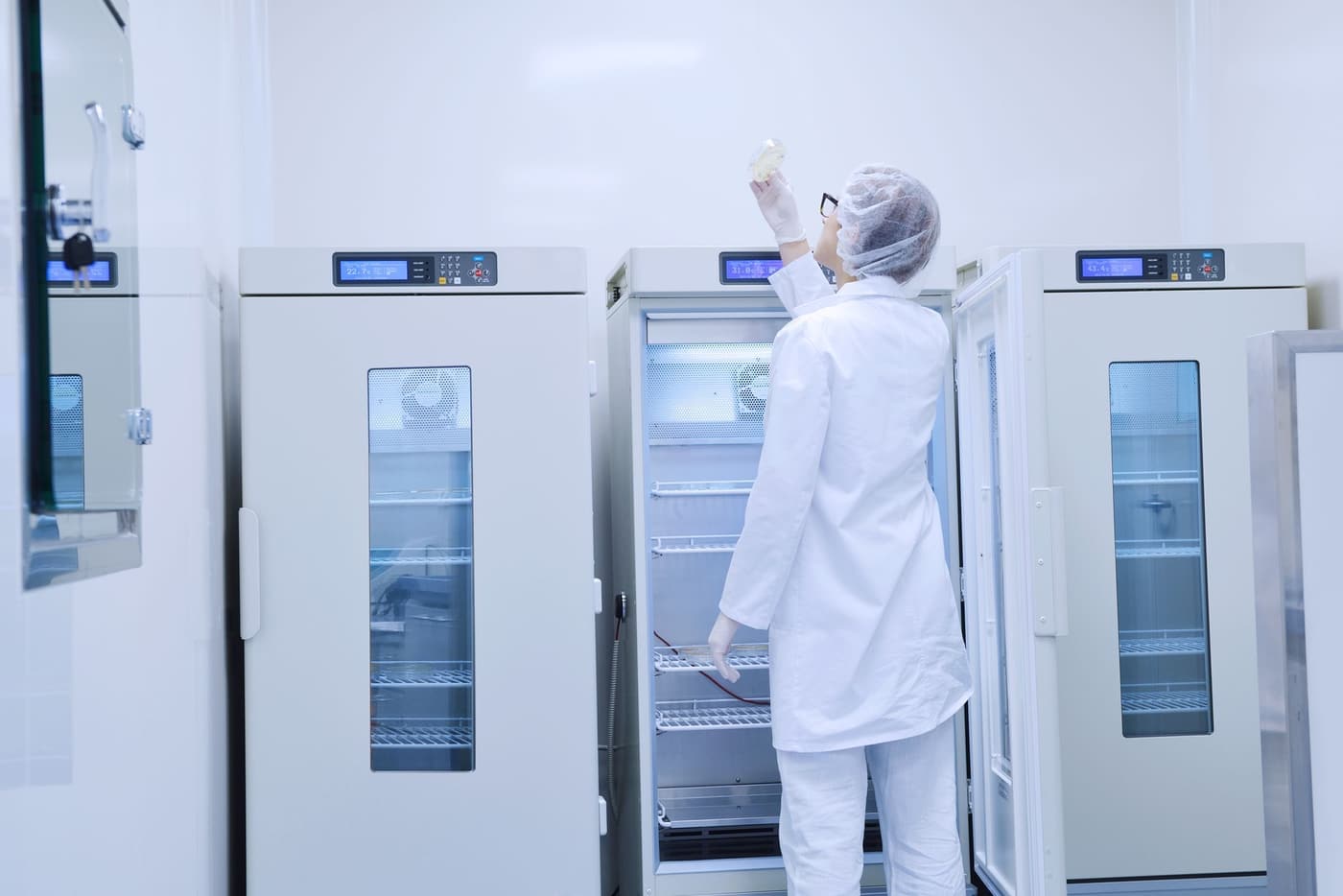 A person in a white lab coat and hairnet places an item inside a tall industrial refrigerator in a bright laboratory.