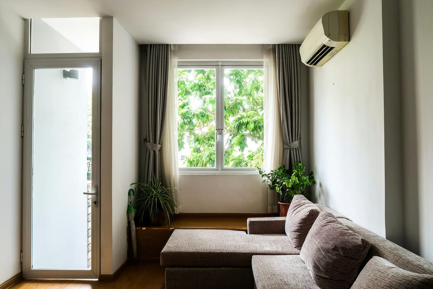 A cozy living room with a gray sectional sofa, a window overlooking trees, sheer and heavy curtains, and an AC unit.